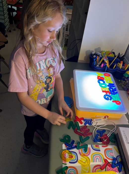 A girl in a pink “PRINCESS” shirt arranges clear plastic letters and numbers on a glowing light box. Letters A–G and numbers 0–8 are visible on the light box. Many loose clear letters and numbers lie on the table.