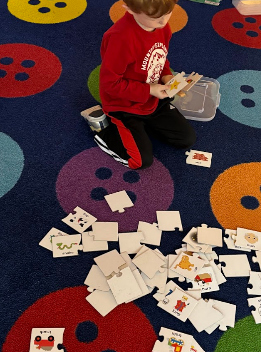 A boy kneels on a blue carpet sorting word-picture puzzle pieces, holding a piece labeled “star.” Scattered pieces show images with words like “snake,” “dog,” “truck,” “train,” “cat,” “barn,” “man,” “box,” and “wall.” A clear plastic container of pieces sits beside him.