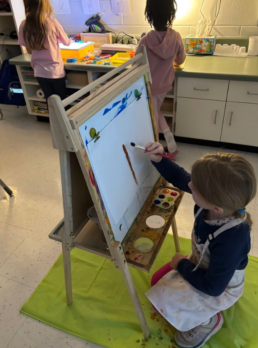 Three children make art; a girl in a paint-splattered apron paints on a small easel while two other children work at a craft table with blocks and supplies. Paint and art materials are visible on the mat and table. The scene shows active, hands-on art exploration.