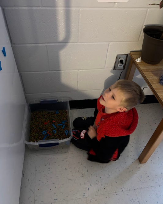 A child in a red hooded jacket sits on a light tile floor smiling and looking up. A clear plastic bin with small colorful beads and blue/red plastic letter shapes sits beside him. A wooden table and cinderblock wall are in the background.