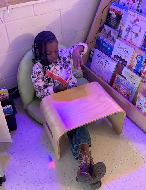 A child sits in a light green beanbag chair reading a red book in a cozy classroom corner. A nearby bookshelf displays children’s books including “Watch Out!,” “Snowmen at Night,” “Little Leaders: Bold Women in Black History,” and “The Day You Begin.” The child appears focused on the book.