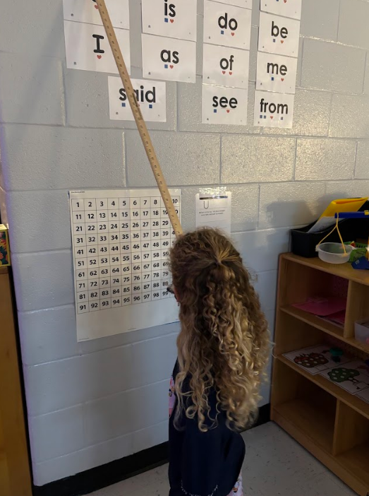 A child points with a wooden ruler at a classroom number chart while sight-word cards above read words like “is,” “do,” “be,” “I,” “as,” “of,” “me,” “said,” “see,” and “from.” Each sight-word card shows two small heart symbols below. Shelving with classroom materials is nearby.