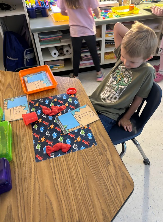 A child wearing a shirt that reads “OFF THE GRID” works with handwriting practice cards. Visible cards say “Handwriting Letters” and show the letters “F” and a stylized “J.” The child is focused at a table.