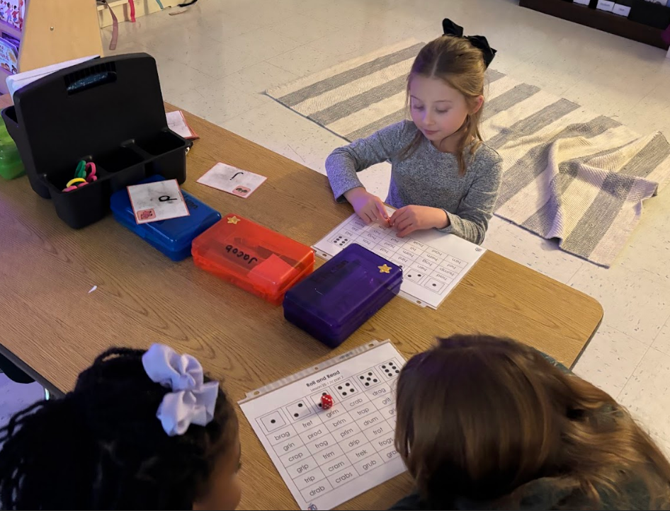 Overhead view of two girls at a table doing “Roll and Read” worksheets; one worksheet shows H-words like “Hug, Ham, Hot,” and the other shows blends like “brag, prod, crab.” Pencil boxes labeled “a” and “Jacob” and a white card with “J” are on the table. A red die rests on one worksheet.