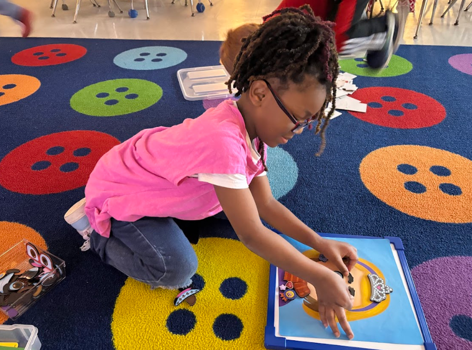 A girl with glasses kneels on a blue carpet assembling a face on a magnetic board using small magnetic pieces. A clear plastic container of pieces is next to her. Classroom furniture legs are visible in the background.