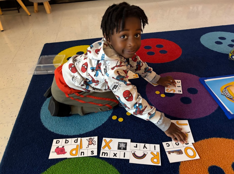 White letter-sound cards are spread on a blue rug with letters and small images (e.g., “p b d” and a pig; “I y r” and a rainbow). A child’s hand reaches toward the cards. Some cards are partially obscured.