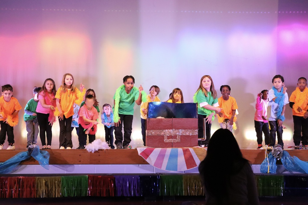 A group of about 15 elementary‑age children perform on a wooden stage lit with colorful spotlights in pink, orange, and purple. They wear bright solid t‑shirts (orange, pink, green, yellow, light blue) with dark pants and fluffy feather boas in matching colors. Children stand across the stage—some smiling, some posing or mid‑movement. Center‑right is a large red prop box with a sparkly gold band and a small blue panel showing a starry scene; a child in yellow stands behind it. The stage front is draped with light blue sheer fabric and white fluff, and a rainbow fringe decorates the lower edge. In the lower right foreground the back of an audience member’s head is visible.