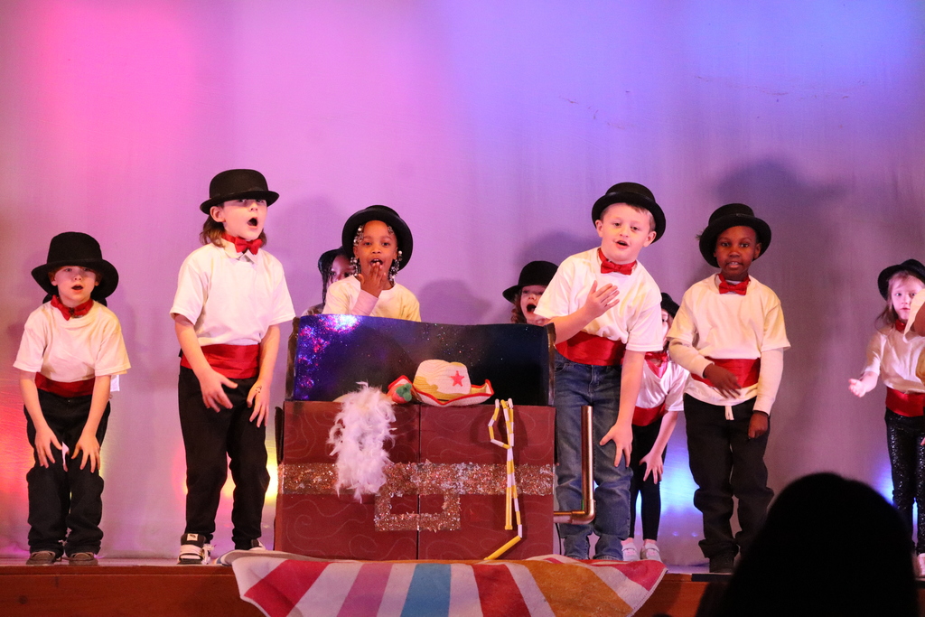 A lively children’s stage performance under purple and pink lighting. Eight+ kids wear matching costumes: black bowler hats, white shirts, red bow ties and red waist sashes, with a mix of dark trousers, jeans and sparkly pants. A large reddish-brown trunk prop with gold accents sits center stage topped with a light hat, a white feathery boa and yellow ribbons. Children around the prop sing and pose—some with hands to their mouths, some leaning forward—conveying energetic, theatrical engagement.