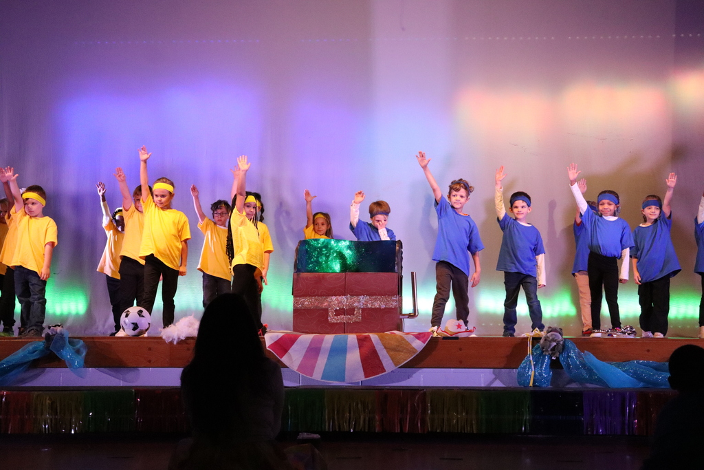 A lively children's stage performance in an auditorium lit with purple, blue and warm accent lights. About 15–20 kids stand across the wooden stage in two color groups: children on the left wear bright yellow t‑shirts and headbands, many with arms raised; children on the right wear blue t‑shirts and headbands with arms raised. A large brown chest‑like prop with a glittery silver band sits center stage, its open top glowing green and blue; one child in yellow and one in blue peer into it. A black‑and‑white soccer ball rests near the left group. The stage front is decorated with colorful striped fabric and reflective bands; a small gray plush sits on iridescent blue fabric at far right. In the lower foreground the backs of audience members’ heads are visible.