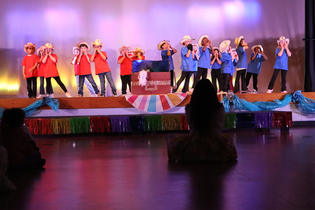 Children perform on a raised stage in a dimly lit hall under purple and pink wash lighting. About 16–18 young performers wear light cowboy hats with star emblems; the left group wears red shirts and the right group wears blue shirts (one child in blue has black gloves). Many children cup their hands around their mouths as if singing or calling. A decorated reddish-brown chest with a white fluffy boa sits center stage, and a colorful fan‑striped fabric drapes the stage front. Metallic rainbow fringe and blue sheer fabric trim the stage edge. Two audience members sit on the floor in the foreground, backs to camera