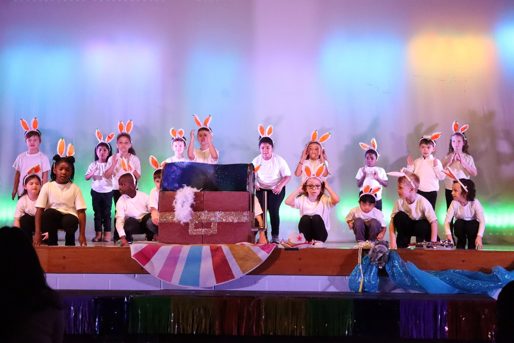 About 18–20 young children perform on a wooden stage under soft pastel lighting. Most wear white t‑shirts and dark bottoms and bright “carrot” headbands with two orange carrot‑shaped attachments. Children are arranged in rows—some standing, some crouching—around a large ornate chest prop with glittery gold bands and a starry dark blue top; a white fluffy decoration is attached to the chest. The stage front is trimmed with colorful fringe and light blue draping; a small stuffed animal and a white hat sit near the right front. The children look engaged and cheerful, facing the audience.