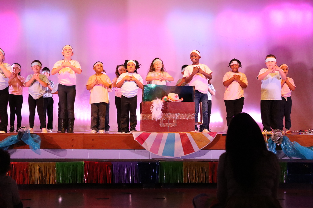 Children perform on a raised wooden stage bathed in pink and purple light. About 10–12 elementary‑age kids stand in a line wearing light t‑shirts, dark bottoms and white headbands, many with hands crossed over their chests in synchronized poses. A glittery red chest prop with a fluffy white decoration and a small pink hat sits center stage, and a blue‑green backdrop peeks behind it. The stage front is trimmed with colorful striped fabric and blue draping. In the lower foreground the back of an audience member’s head is visible