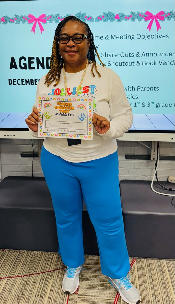 A smiling kindergarten teacher stands in a classroom holding a colorful “Teacher Character Trait Award for Citizenship” certificate, with a presentation screen behind her.