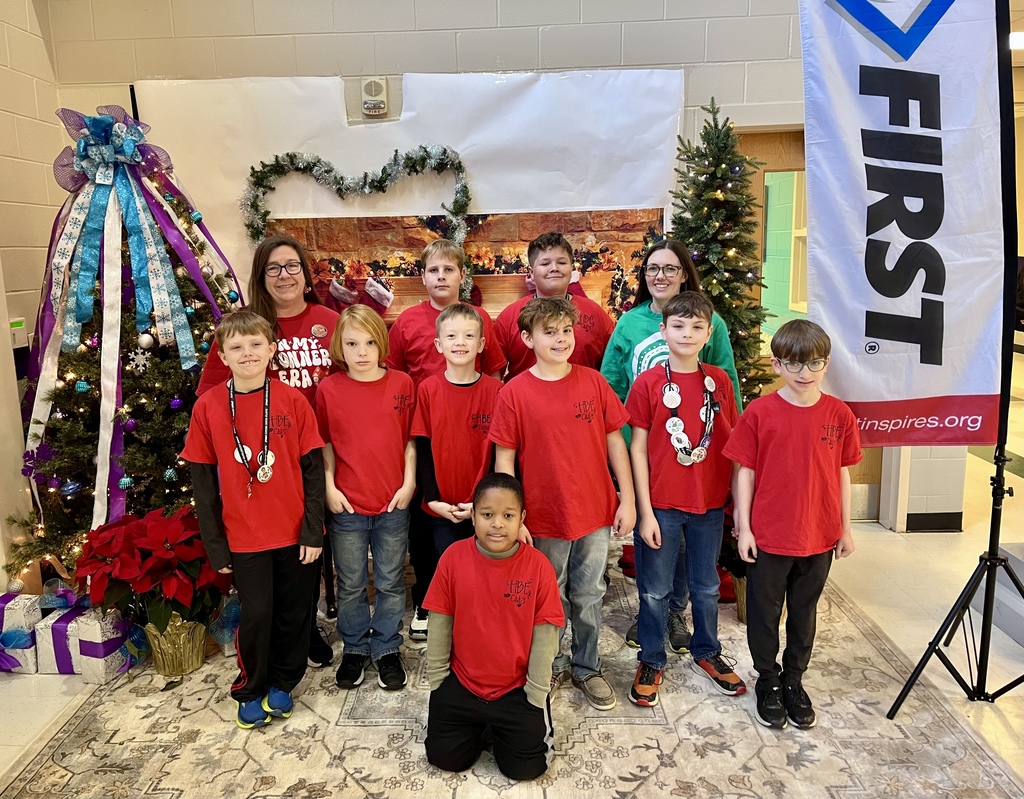 Photo of an indoor event showing several children in the foreground and midground. Kids of varying ages stand and sit, some looking toward the camera and others focused on activities off-frame. They wear casual clothing and appear engaged and curious—one child leans forward attentively while another smiles. In the center background, an adult woman in a red shirt. To the right, a vertical banner shows the FIRST logo and the URL “tinspires.org.” 