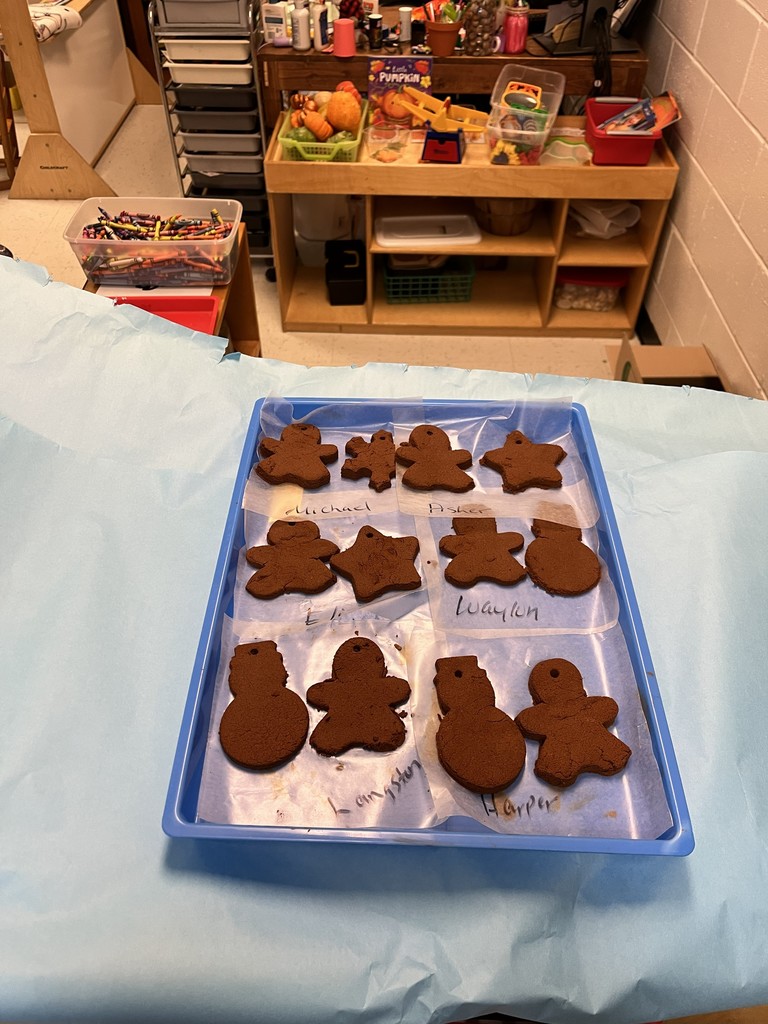 Classroom scene showing a blue tablecloth with a tray of unbaked gingerbread cookies in rows — shapes include gingerbread people, stars, and snowmen. Each cookie rests on a small square of parchment with a handwritten name underneath