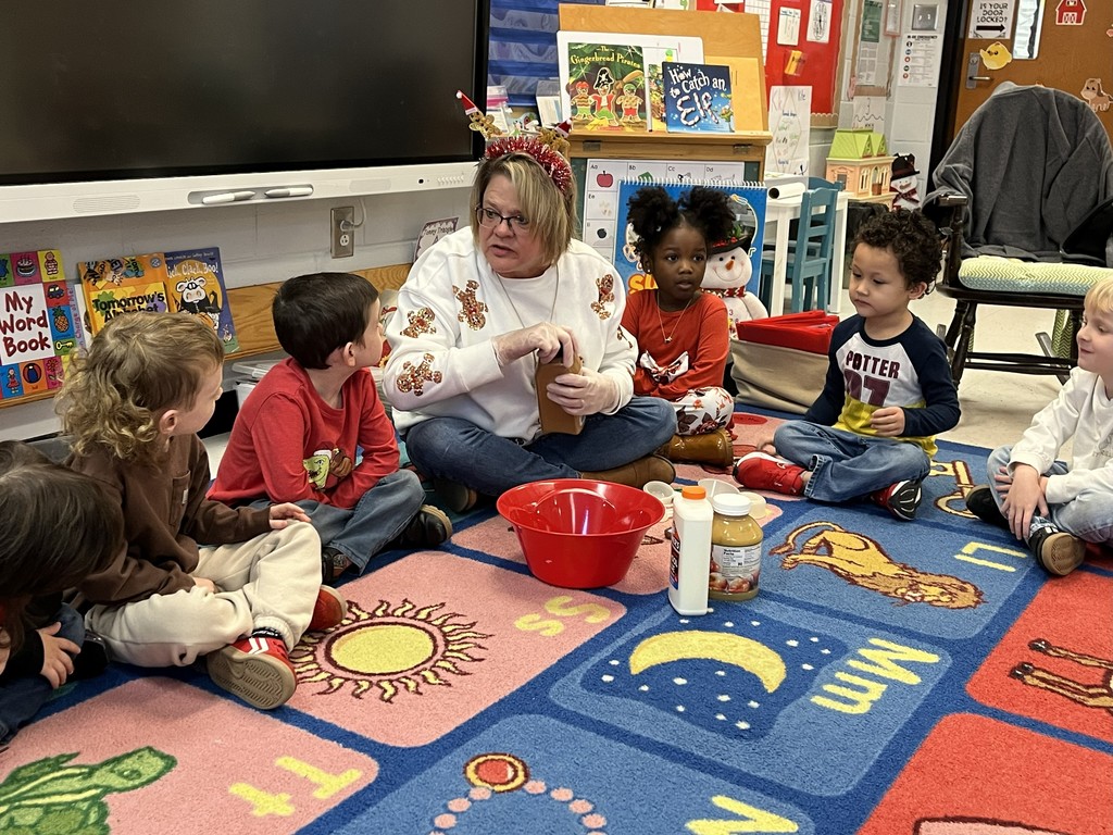 Classroom scene on a colorful alphabet rug: an adult woman in a white sweater patterned with gingerbread men and wearing reindeer antler headband sits with six young children gathered around her. She holds a brown container and wears clear gloves while demonstrating an activity; nearby are a red mixing bowl, a bottle of glue, and a jar of applesauce. Children watch attentively