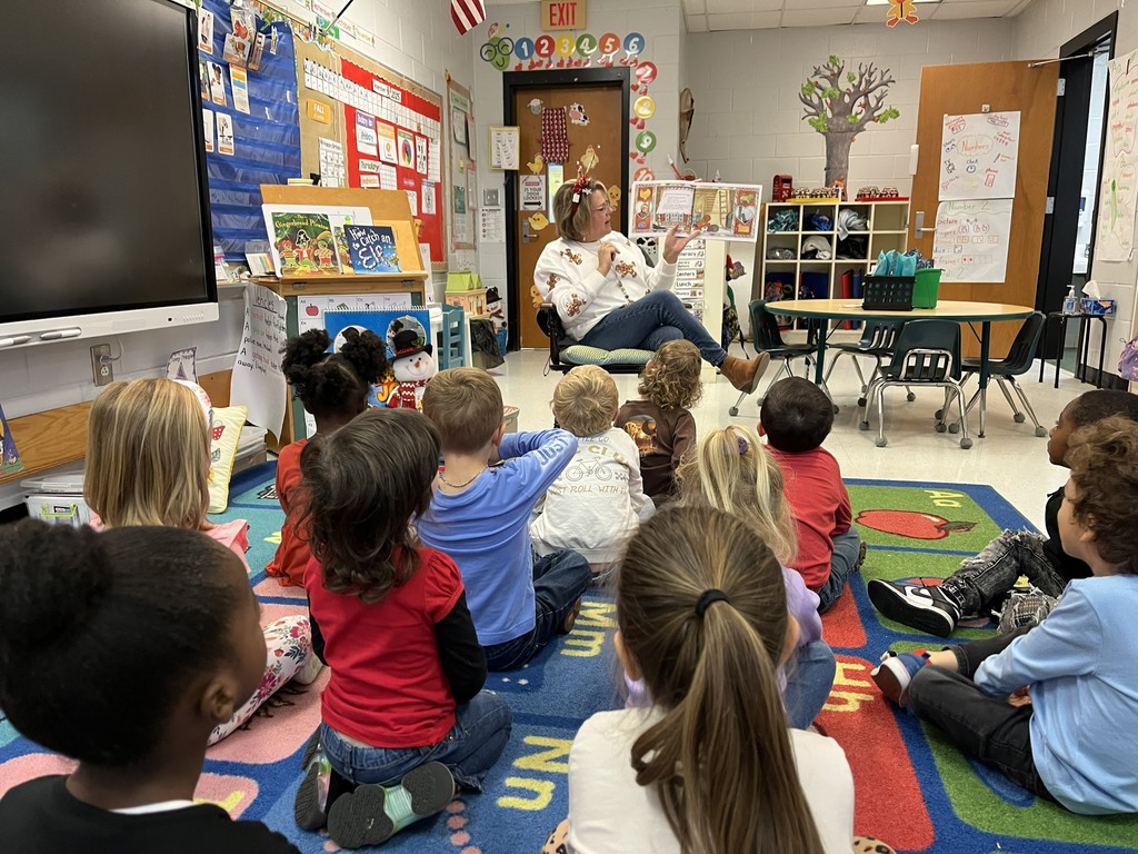 Teacher reading to a group of about 10–12 young children seated on a colorful blue rug. The teacher wears a white shirt with gingerbread cookie designs, jeans, and a festive headband, and holds an open book.