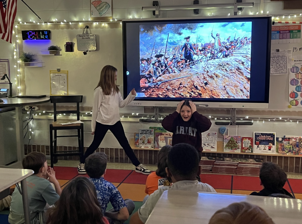 Two girls stand in front of a large screen displaying a historical battle painting; one gestures dramatically while classmates sit on a red carpet watching. Books with holiday themes line the shelf beneath the screen.