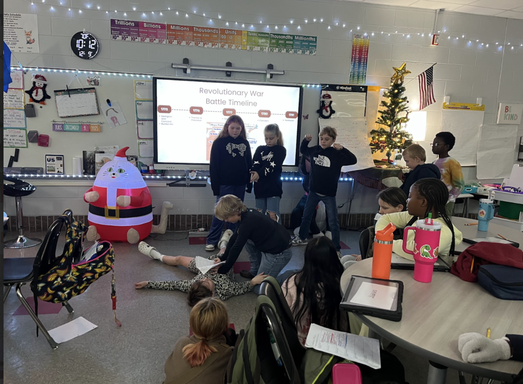 (Revolutionary War timeline): Classroom with a large screen showing a Revolutionary War battle timeline (1775–1781) listing fights like Lexington & Concord and Bunker Hill. Three students stand near the screen while others sit at round tables or on the floor. Holiday decorations (string lights, small lit tree) and an inflatable Santa‑cat are visible.