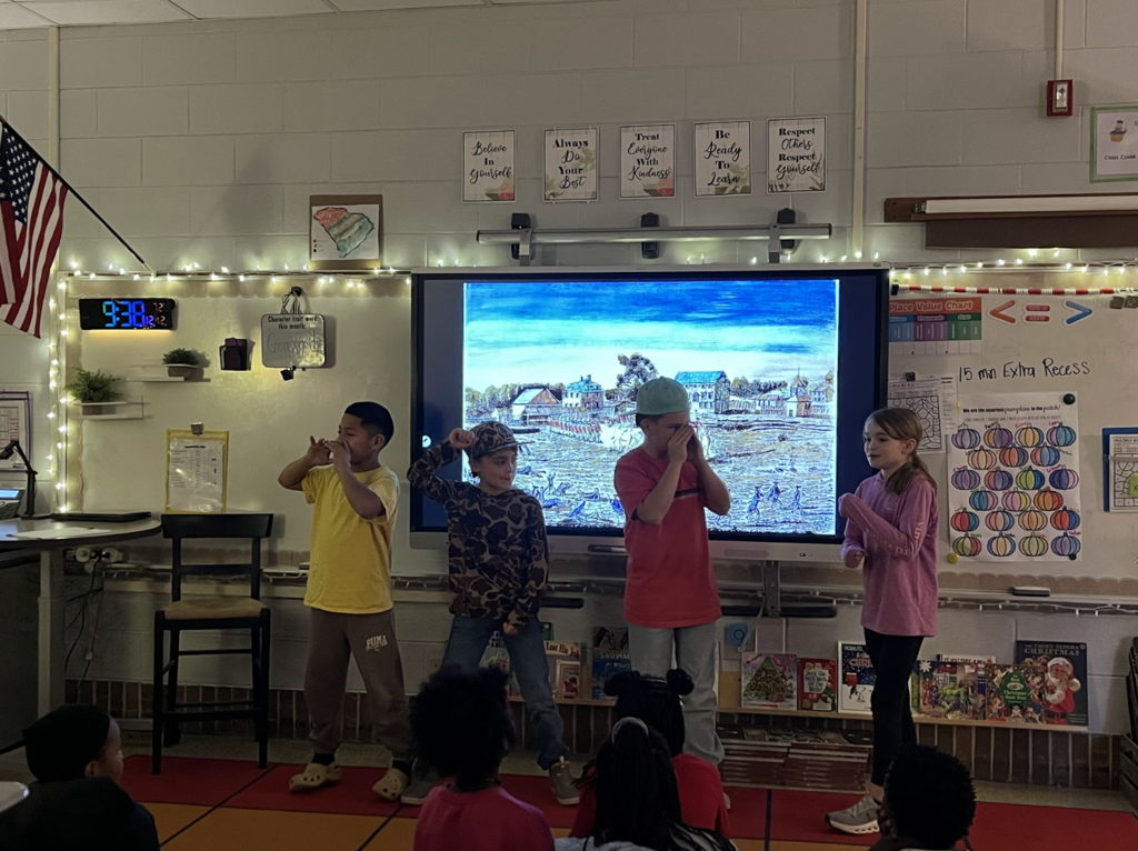 Four children stand and make hand gestures in front of an interactive display while peers watch from the carpet