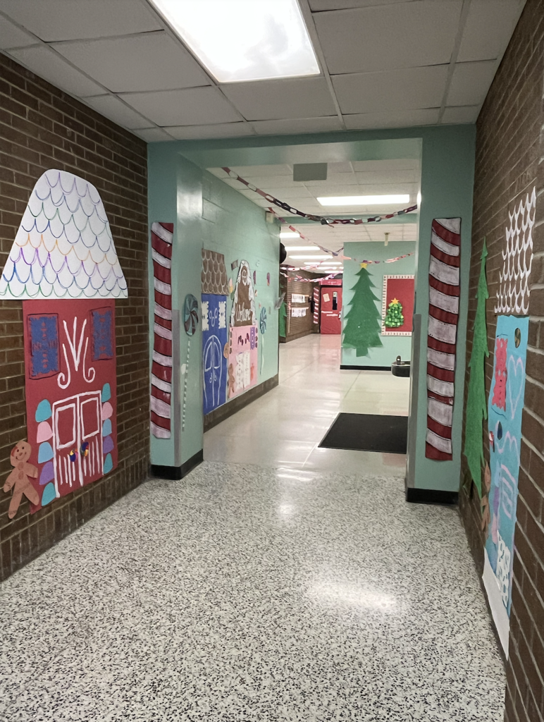 School hallway decorated like a gingerbread village with brown brick and mint-green walls, large red and colorful gingerbread house cutouts, candy‑cane columns, a paper‑chain garland overhead, and a green Christmas tree cutout on the right
