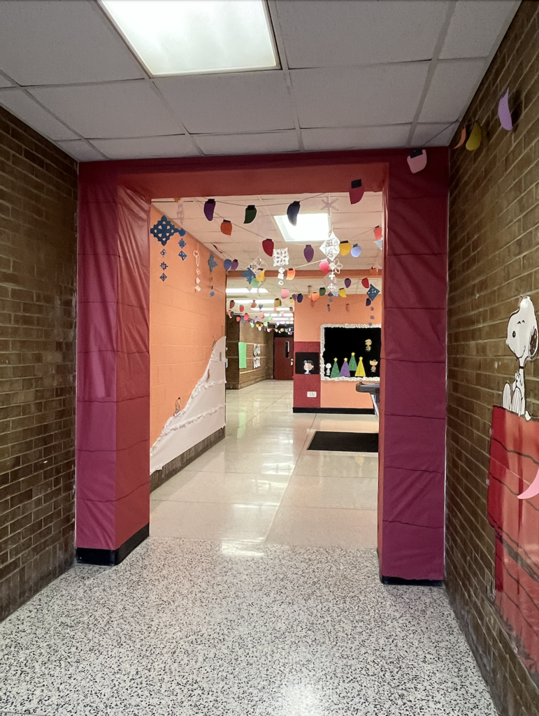 Hallway framed by a dark-pink fabric-wrapped archway leading to a decorated inner corridor; Snoopy-in-doghouse cutout on the right, hanging paper lightbulb decorations, paper snowflakes from the ceiling, and colorful wall displays.