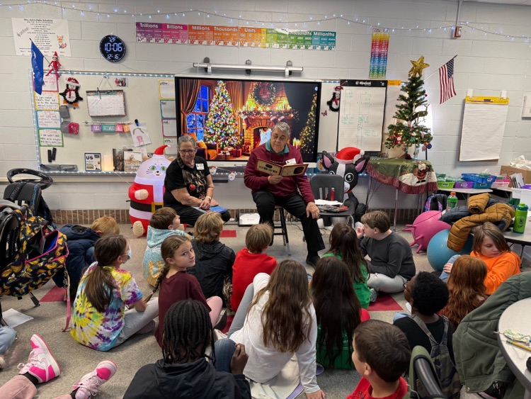 Elementary students sit on the floor listening to two guest readers in a classroom decorated for the holidays.
