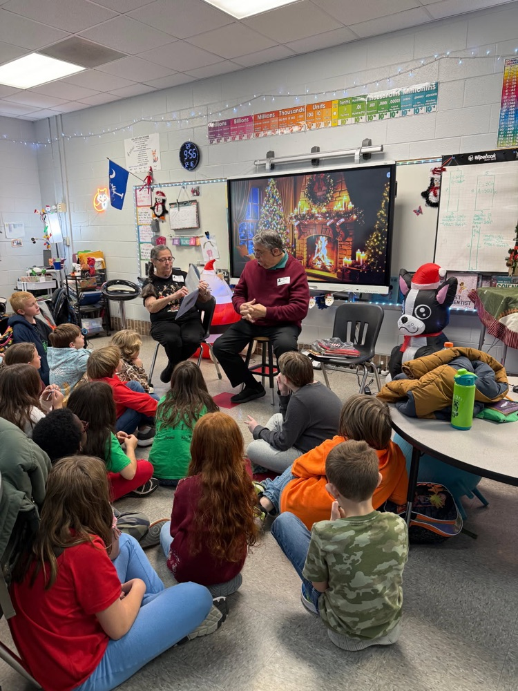 Elementary students sit on the floor listening to two guest readers in a classroom decorated for the holidays.