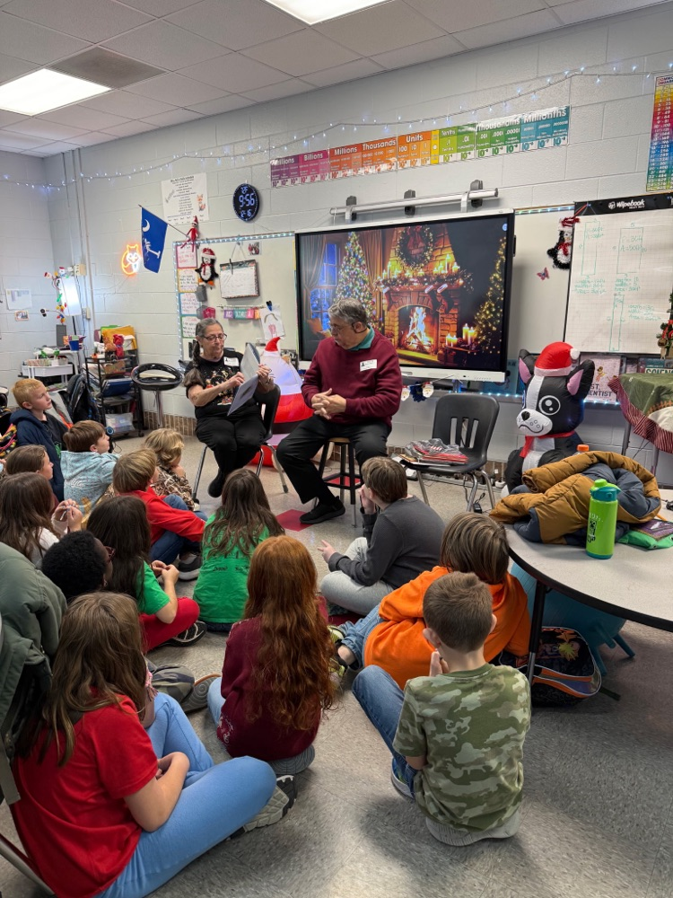 Elementary students sit on the floor listening to two guest readers in a classroom decorated for the holidays.
