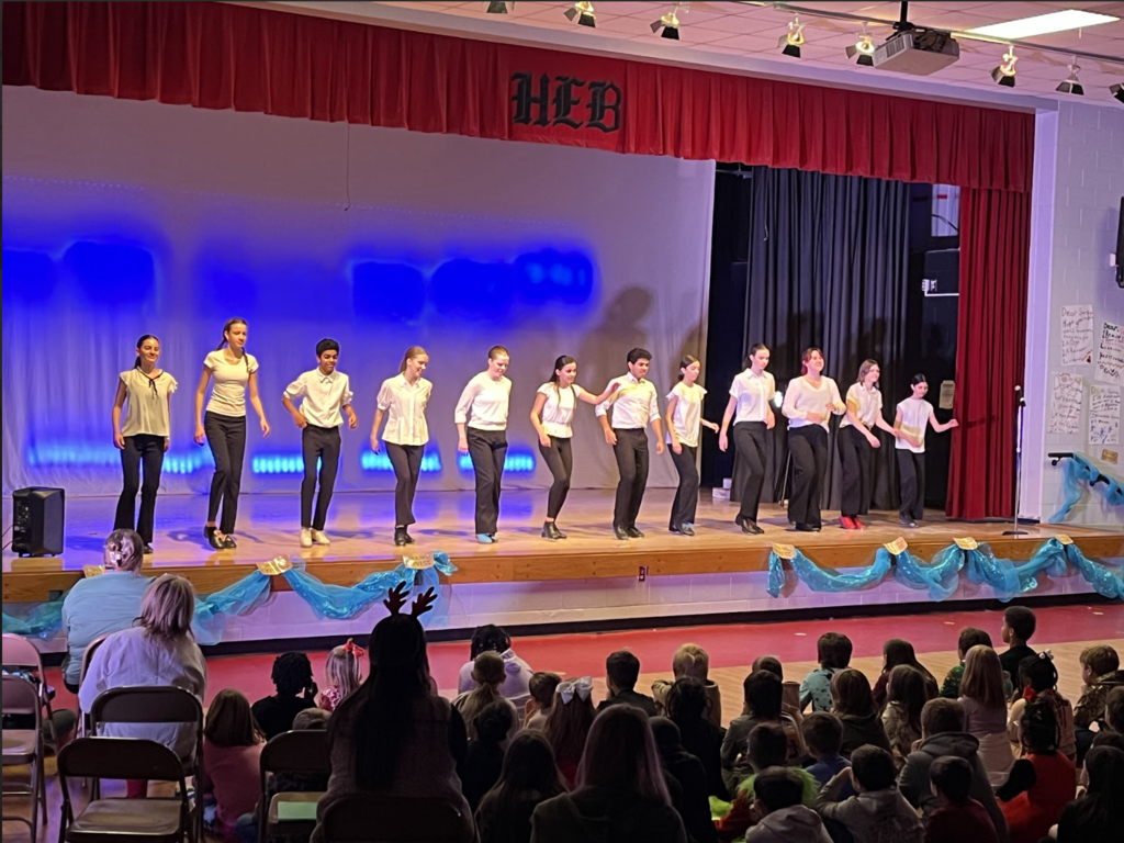 Indoor school performance in a gym/multipurpose room. A stage with red curtains and a blue‑lit backdrop holds about 8–10 young performers in light tops and dark bottoms, mid‑movement in a line. The front of the stage is decorated with light blue fabric and gold accents. In the foreground a large audience—mostly children seated on the floor with adults in chairs behind them—watches. 3