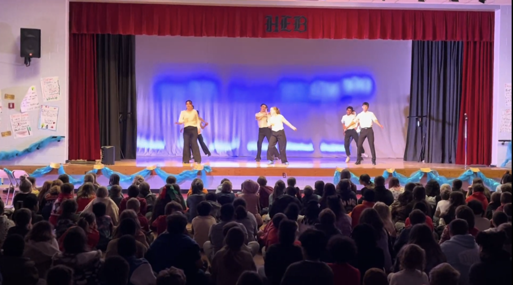 Indoor school performance in a gym/multipurpose room. A stage with red curtains and a blue‑lit backdrop holds about 8–10 young performers in light tops and dark bottoms, mid‑movement in a line. The front of the stage is decorated with light blue fabric and gold accents. In the foreground a large audience—mostly children seated on the floor with adults in chairs behind them—watches. 2