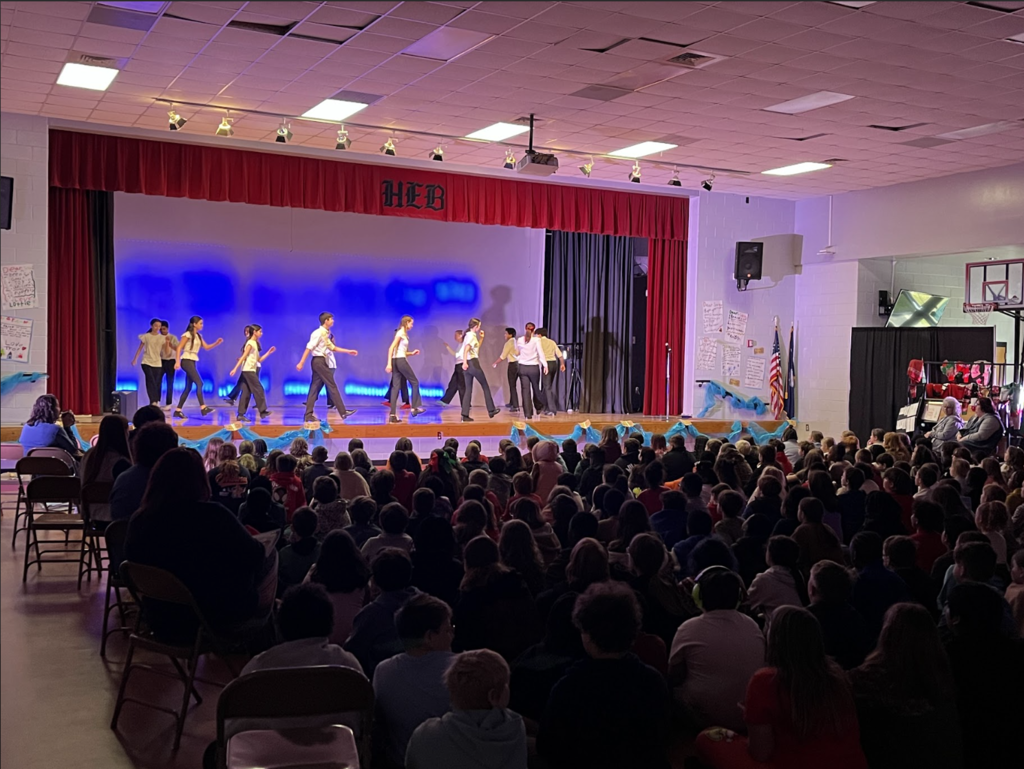 Indoor school performance in a gym/multipurpose room. A stage with red curtains and a blue‑lit backdrop holds about 8–10 young performers in light tops and dark bottoms, mid‑movement in a line. The front of the stage is decorated with light blue fabric and gold accents. In the foreground a large audience—mostly children seated on the floor with adults in chairs behind them—watches. 4