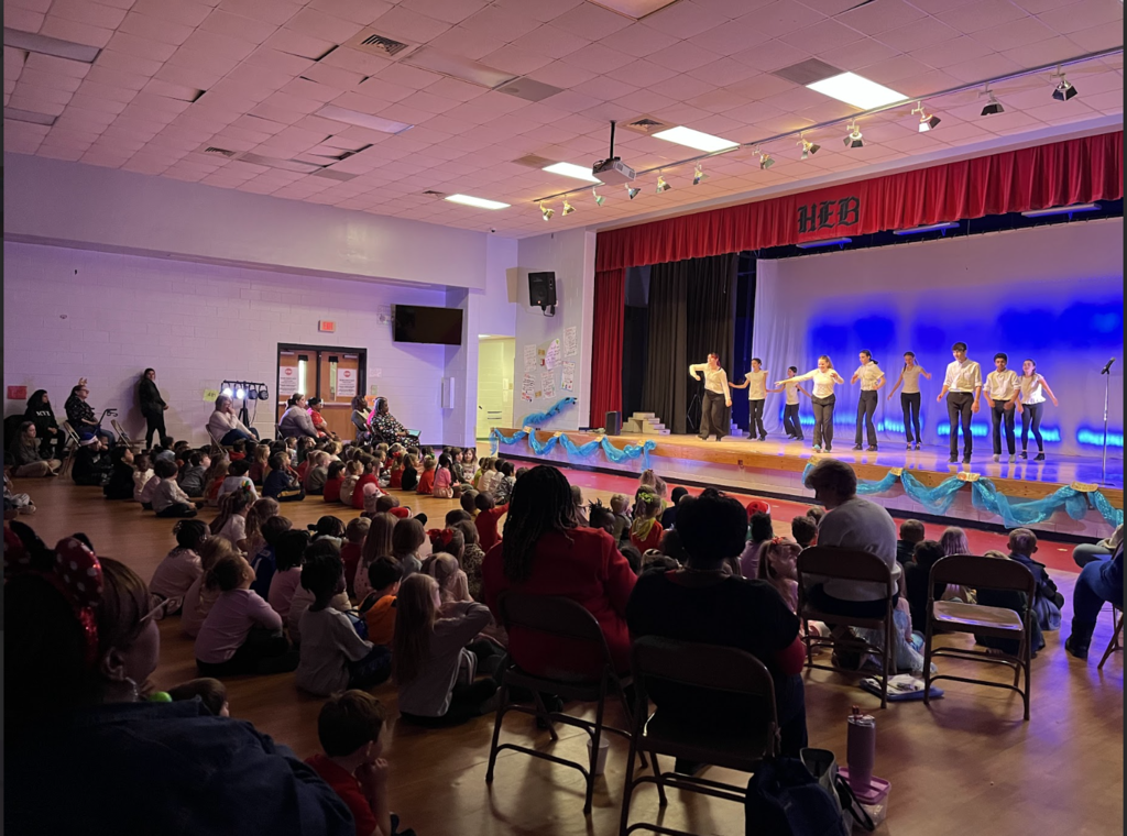 Indoor school performance in a gym/multipurpose room. A stage with red curtains and a blue‑lit backdrop holds about 8–10 young performers in light tops and dark bottoms, mid‑movement in a line. The front of the stage is decorated with light blue fabric and gold accents. In the foreground a large audience—mostly children seated on the floor with adults in chairs behind them—watches. 1