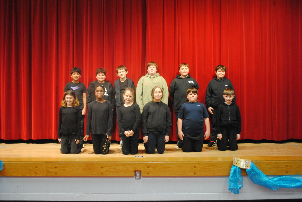 Picture features 15 students in all black in front of a red curtain. 