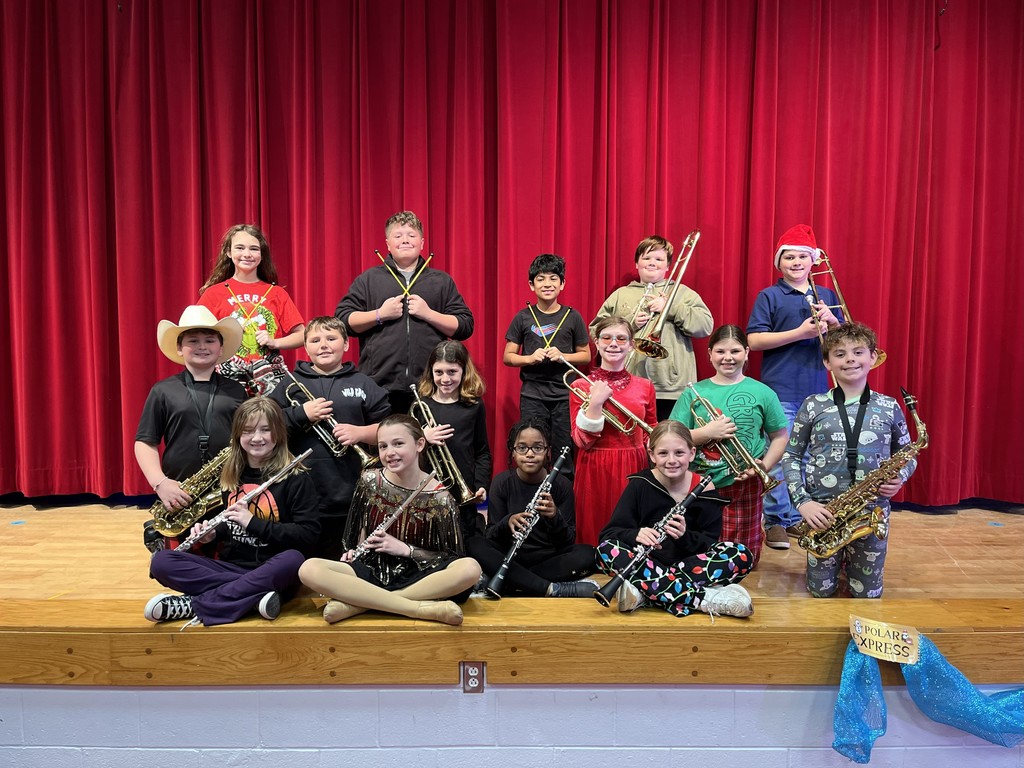 15 students holding band instruments (Flute, clarinet, saxophone, trumpet, trombone, percussion mallets) in front of a red curtain wearing holiday gear.