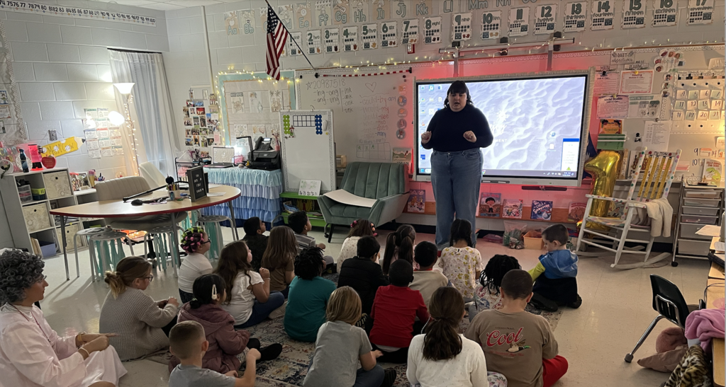First grade students in their classroom with artist Bailey Mann! Students are sitting on carpet while artist is in front of smart board. 
