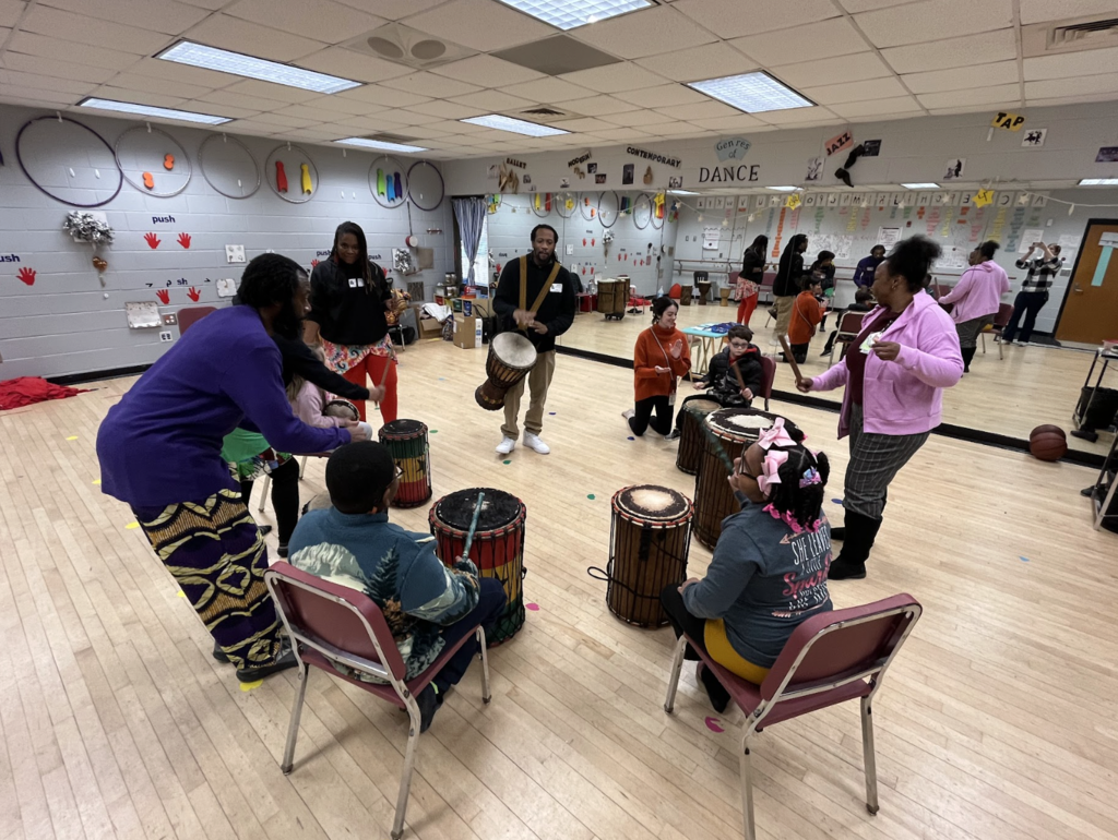 Students sitting in a circle with artists around them holding drums and playing their instrument