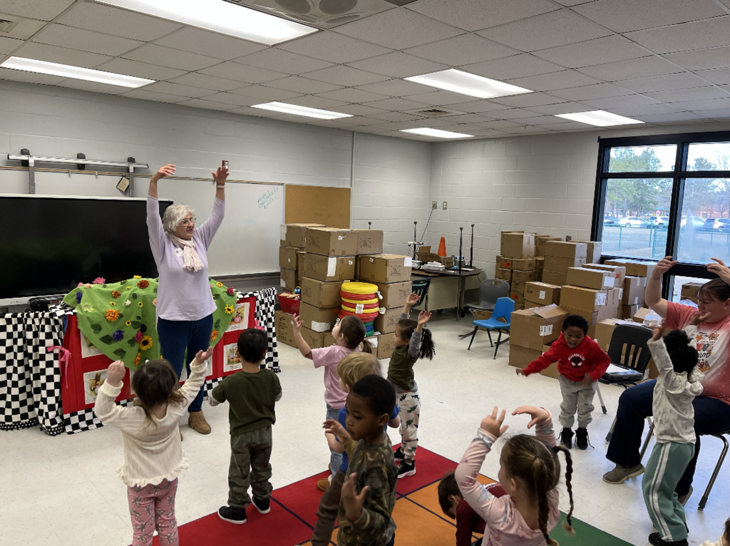Teacher leads prek students on a colorful rug; woman with grey hair raises her arms as children mimic her. Whiteboard reads “WELCOME!! Class of ’27!!”; stacked boxes and windows visible
