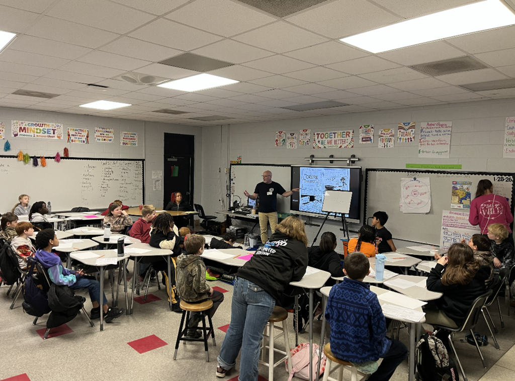 Students sit at desks in a classroom while a teaching artist stands at the front, presenting a lesson on a digital board. Students watch and listen as the instructor gestures and explains the activity.