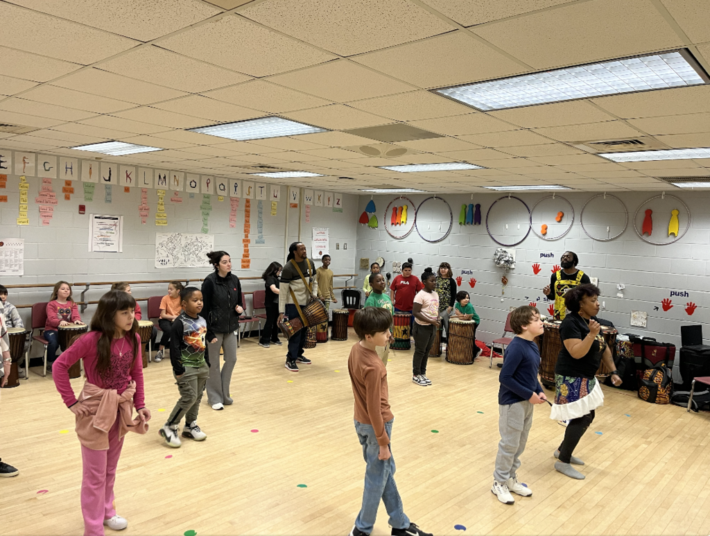 Elementary students stand and move in a music or dance classroom while teaching artists play drums. Students follow along with movement as part of an interactive music and dance lesson.