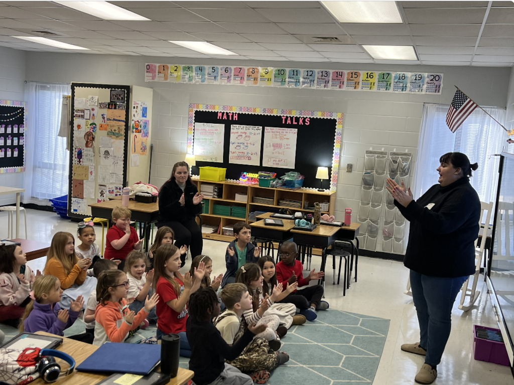 Students sitting on a blue carpet clapping their hands with teacher in front of classroom