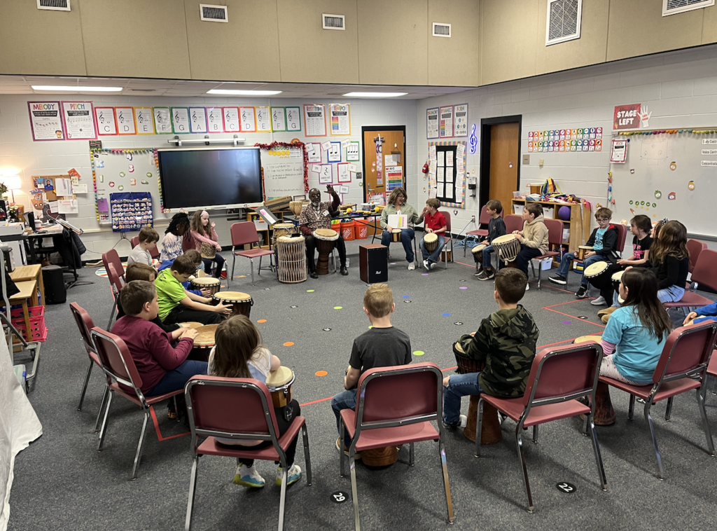 Students sitting in a circle on chairs playing djembes 