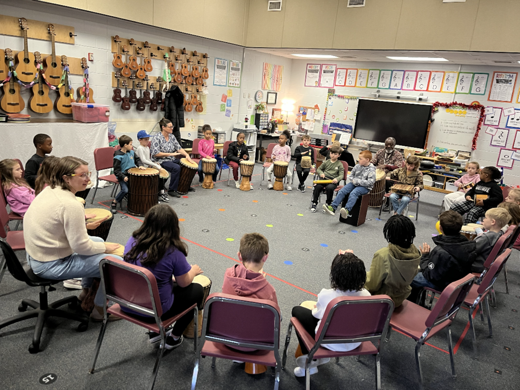 Students sitting in a circle in chairs playing a variety of drums. 