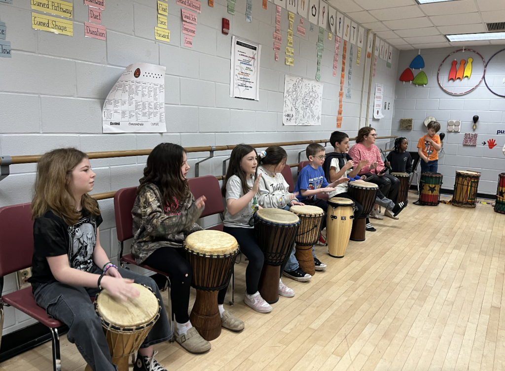 Students lining the wall in chairs playing a variety of djembes 