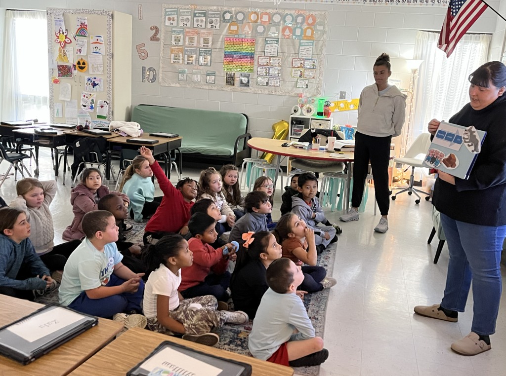 Students sitting on a carpet creating a new ending to the three little pigs, a story board is being held by the teacher 