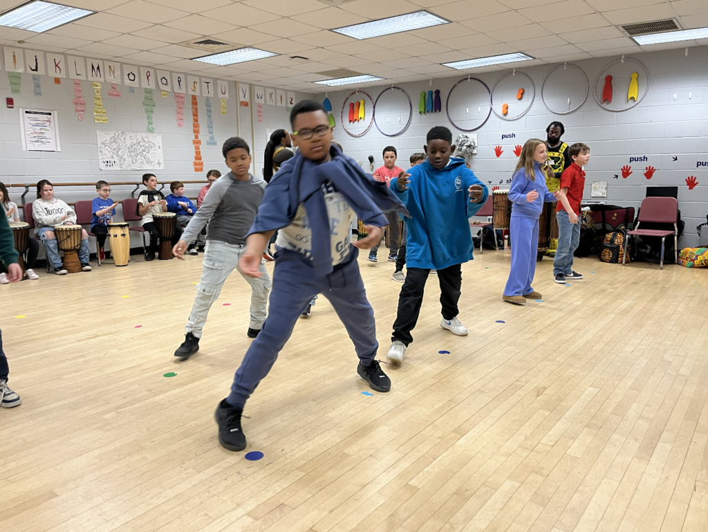 Students dancing in the dance room, drums in the background, students are moving arms froward and feet to the side 