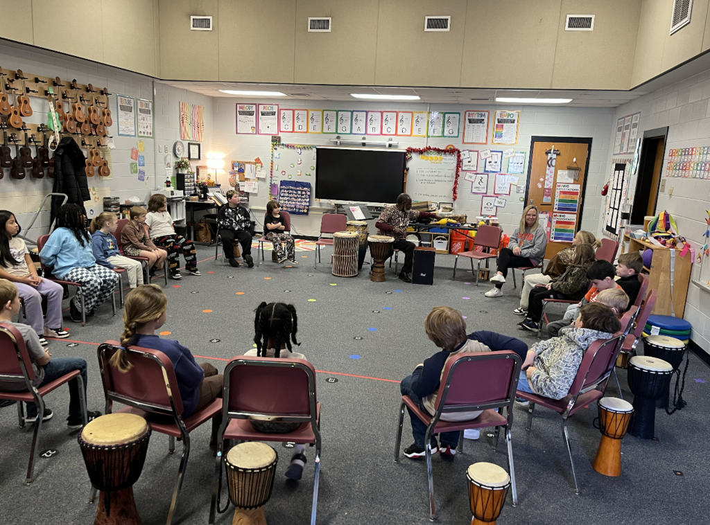 Students sitting in the music room on chairs with djembes! Instructor is showing a variety of instruments from Africa 