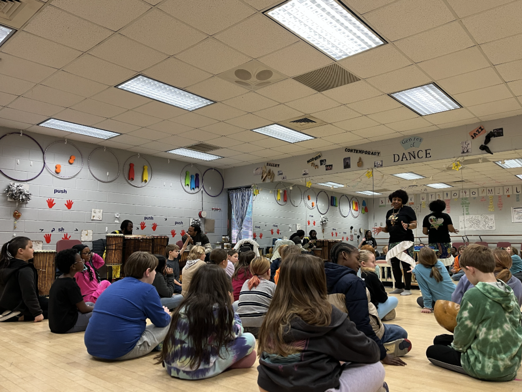 Students sitting on the floor looking up a the artist standing in front of the mirror.
