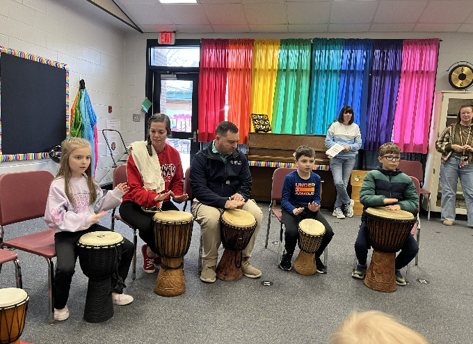 3 children and 2 adults sitting at djembes, rainbow curtains in are in the background 