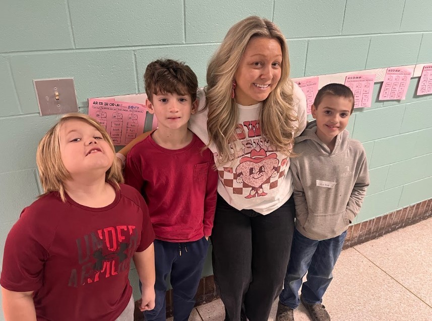 Four people standing against a light blue-green brick wall: two boys at left (one in dark red Under Armour shirt, one in red long-sleeve), a smiling woman in a white T-shirt with a pink-checkered heart graphic, and a smiling boy at right wearing a gray hoodie with a name tag reading 'Kai Day'. Pink educational sheets on the wall show 'AR ER IR OR UR' and a list of words.